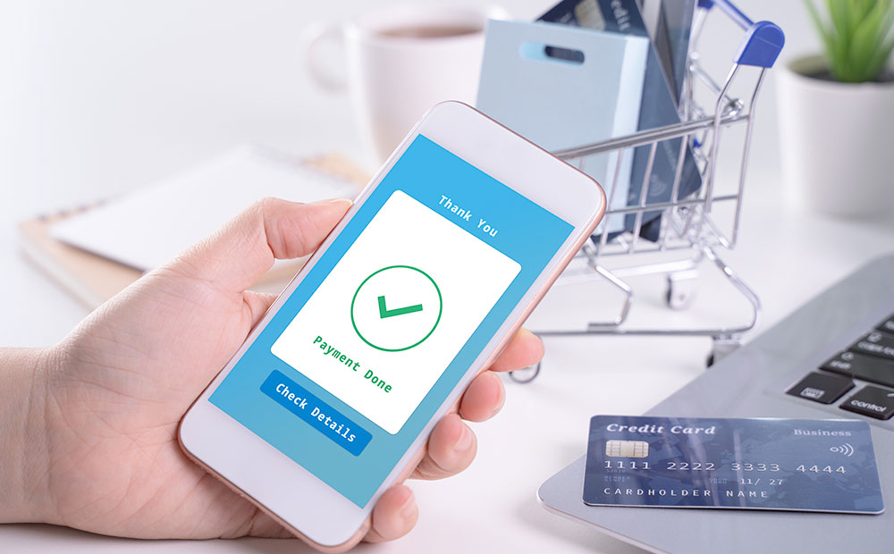 ekranında "thank you. payment done. check details" yazılı akıllı telefonu tutan insan eli ve üzerinde kredi kartı ve laptop bulunan masa fotoğrafı